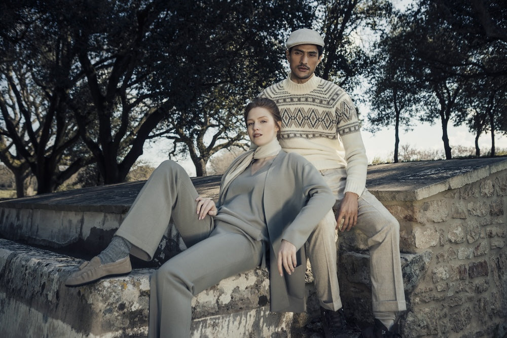 Woman wearing a grey outfit and man in a light patterned jumper, sitting on a stone wall outdoors.